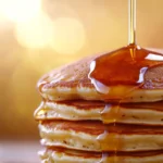 A stack of golden-brown pancakes with crispy edges topped with maple syrup dripping over the sides, illustrating How to Keep Pancakes Crispy?, glowing against a warm, golden background.